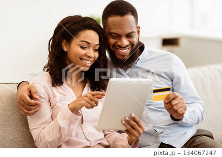 Online Shopping And Ecommerce. Happy young african american family of two people sitting on sofa at home, choosing and buying clothes or products online, paying with credit card using digital tablet 134067247