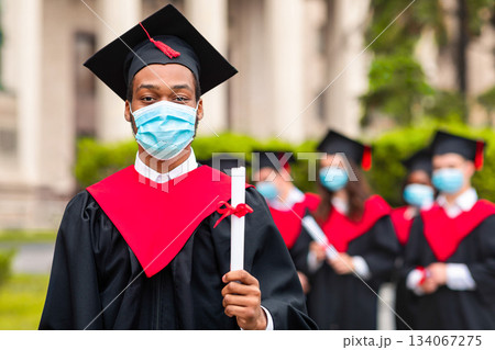 Black guy student in graduation costume showing diploma at camera, posing over international group of students at university campus, wearing protective face mask, panorama with copy space 134067275