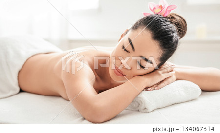 Spa Relaxation. Young Asian Woman Lying On Massage Table In Beauty Salon, Calm Korean Lady With Exotic Flower In Hair Resting Before Body Treatment, Closeup Shot, Free Space Spa Relaxation. Young Asian Woman Lying On Massage Table In Beauty Salon, Calm Korean Lady With Exotic Flower In Hair Resting Before Body Treatment, Closeup Shot, Free Space 134067314
