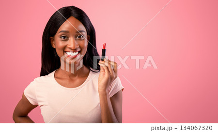 Attractive black lady holding lipstick over pink studio background. Millennial African American woman holding tube of makeup, applying cosmetic product, promoting lip gloss 134067320