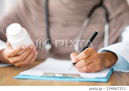 Medicine, pills, vitamins concept. Closeup of unrecognizable female doctor in hijab holding medicine, writing prescription in medical chart, prescribing treatment for patient in hospital, cropped Medicine, pills, vitamins concept. Closeup of unrecognizable female doctor in hijab holding medicine, writing prescription in medical chart, prescribing treatment for patient in hospital, cropped 134067351
