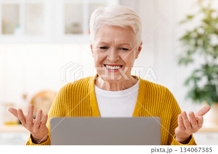 Happy elegant old woman sitting in kitchen, using modern laptop and gesturing, having video call with family, attending video conference or webinar, drinking tea and taking notes, copy space 134067385
