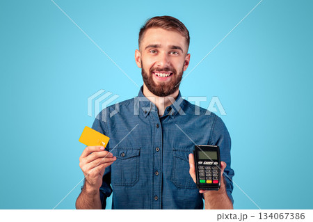 Portrait of happy young guy holding credit card and payment terminal, amiling and looking at camera over blue studio background. Positive millennial man shopping contactless during covid outbreak 134067386