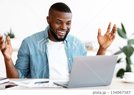 Happy Black Freelancer Man Celebrating Success While Working Online On Computer, African Guy Looking At Laptop Screen, Raising Hands And Exclaiming With Excitement, Sitting At Desk In Home Office 134067387