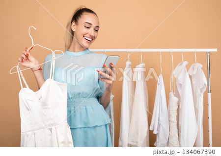 Portrait of smiling young woman using her mobile phone, holding hanger with dress, chatting and sending photos to her friend, choosing outfit, standing near clothing rail at the studio 134067390