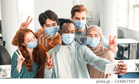 Friendship, Lifestyle And People Concept. Portrait of smiling happy diverse multiracial group wearing surgical face masks and taking selfie indoors, using mobile phone, showing peace or victory sign Friendship, Lifestyle And People Concept. Portrait of smiling happy diverse multiracial group wearing surgical face masks and taking selfie indoors, using mobile phone, showing peace or victory sign 134067397