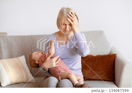 Exhausted blonde woman young mother having migraine, holding crying little baby, home interior, copy space. Tired caucasian mom having headache, touching her forehead. Stressful motherhood concept Exhausted blonde woman young mother having migraine, holding crying little baby, home interior, copy space. Tired caucasian mom having headache, touching her forehead. Stressful motherhood concept 134067399