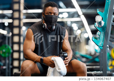 Sport during epidemic, safety first. African american athletic man with wireless headphones wearing protective mask while exercising at gym, holding protein drink and towel, break time, free space 134067407