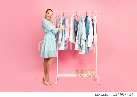 Full length vertical portrait of cheerful smiling woman standing at boutique wardrobe, choosing fashion stylish clothes at rack with hangers, looking at camera, looking for outfit, pink background 134067414