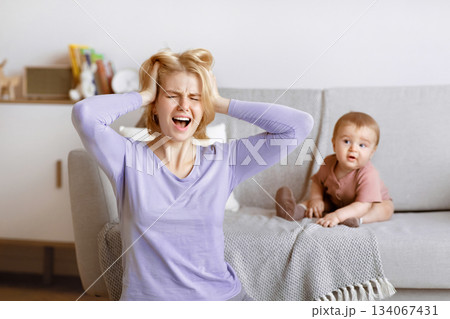 Postnatal depression concept. Stressed blonde woman young mother having headache with baby at home, sitting on floor, touching her head and screaming. Motherhood, multi-tasking and family 134067431