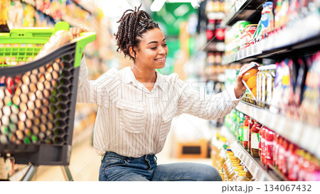 African Woman Buyer Choosing Products Doing Grocery Shopping Buying Healthy Organic Food In Local Supermarket, Posing With Shop Cart Full Of Groceries. Happy Female Customer Concept African Woman Buyer Choosing Products Doing Grocery Shopping Buying Healthy Organic Food In Local Supermarket, Posing With Shop Cart Full Of Groceries. Happy Female Customer Concept 134067432