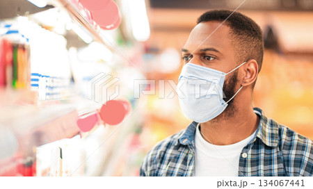 Supermarket Consumer's Portrait. African Man Wearing Face Mask Doing Grocery Shopping Buying Food In Groceries Shop, Standing Near Shelves In Store Aisle Choosing Products Indoors Supermarket Consumer's Portrait. African Man Wearing Face Mask Doing Grocery Shopping Buying Food In Groceries Shop, Standing Near Shelves In Store Aisle Choosing Products Indoors 134067441