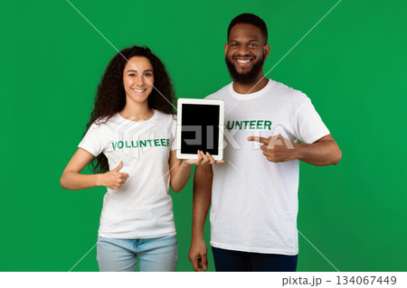 Diverse Volunteers Holding Digital Tablet Pointing At Blank Computer Screen And Gesturing Thumbs Up Standing In Studio On Green Background, Smiling To Camera. Mockup Diverse Volunteers Holding Digital Tablet Pointing At Blank Computer Screen And Gesturing Thumbs Up Standing In Studio On Green Background, Smiling To Camera. Mockup 134067449