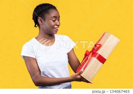 Happy African Lady Holding Wrapped Gift Box Standing Over Yellow Studio Background. Birthday Presents And Gifts, New Year And Christmas Holiday Celebration Concept 134067465