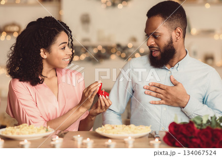 Be My Husband. Smiling black woman making proposal to her surprised man, showing present, holding open box with marriage ring, asking to get engaged during romantic dinner at home or cafe 134067524