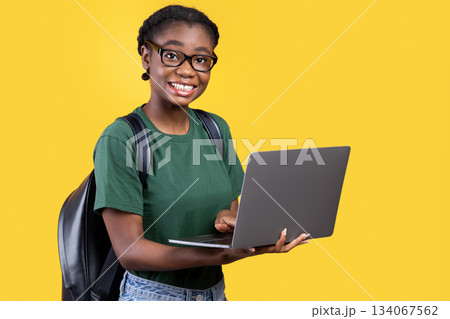 Happy Black Student Girl Using Laptop Computer Posing Smiling To Camera Standing Over Yellow Background. Studio Shot. E-Learning And Online Education. Educational Gadgets Concept 134067562