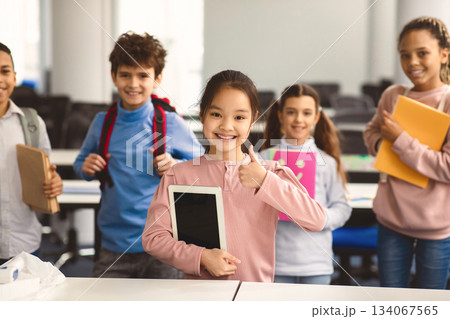 Education And Technology Concept. Smiling small asian girl holding digital tablet and showing thumbs up sign gesture. Diverse group of international classmates standing with backpacks in background 134067565