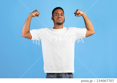 Strength. Strong Black Guy Showing Biceps Looking At Camera Posing Standing On Blue Background In Studio Strength. Strong Black Guy Showing Biceps Looking At Camera Posing Standing On Blue Background In Studio 134067707