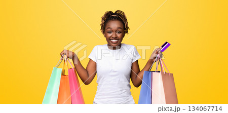 Happy black lady with shopping bags and credit card smiling over yellow studio background, empty space. African american woman shopaholic holding bunch of purchases and unlimited credit card 134067714