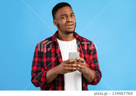 Unsure Black Man Holding Cellphone Posing Over Blue Studio Background. Unpleasant Call Concept Unsure Black Man Holding Cellphone Posing Over Blue Studio Background. Unpleasant Call Concept 134067768