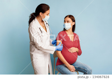 Covid-19 Vaccination. General Practitioner Vaccinating Pregnant Female Patient Against Coronavirus With Antiviral Vaccine, Sticking Medical Adhesive Bandage On Arm After Injection On Blue Background 134067828