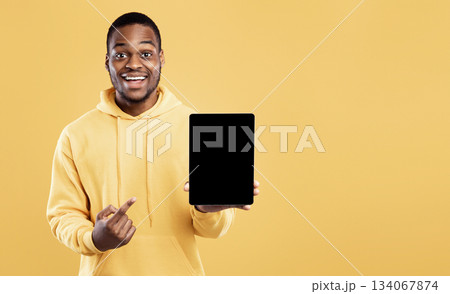 Black Man Pointing At Digital Tablet Empty Screen Holding Computer Posing Over Yellow Studio Background, Smiling To Camera. Mockup For Great Website Or Applicatioin Advertisement Black Man Pointing At Digital Tablet Empty Screen Holding Computer Posing Over Yellow Studio Background, Smiling To Camera. Mockup For Great Website Or Applicatioin Advertisement 134067874
