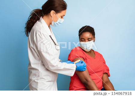 Antiviral Immunization, Covid-19 Vaccination. Black Female Getting Vaccinated, Doctor Sticking Adhesive Bandage After Vaccine Injection On Patient's Arm Over Blue Background. Studio Shot Antiviral Immunization, Covid-19 Vaccination. Black Female Getting Vaccinated, Doctor Sticking Adhesive Bandage After Vaccine Injection On Patient's Arm Over Blue Background. Studio Shot 134067918