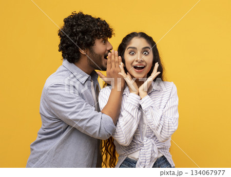Shock, gossip, share advice. Young indian man whispering to lady on ear, happy woman with open mouth excited and surprised, posing on yellow studio background Shock, gossip, share advice. Young indian man whispering to lady on ear, happy woman with open mouth excited and surprised, posing on yellow studio background 134067977