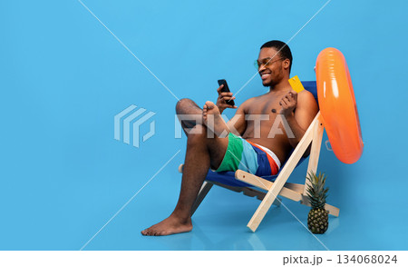 Online summer sale. Carefree black man holding credit card and smartphone, shopping on web in lounge chair, blue background, panorama with empty space. Handsome young guy purchasing goods on internet Online summer sale. Carefree black man holding credit card and smartphone, shopping on web in lounge chair, blue background, panorama with empty space. Handsome young guy purchasing goods on internet 134068024
