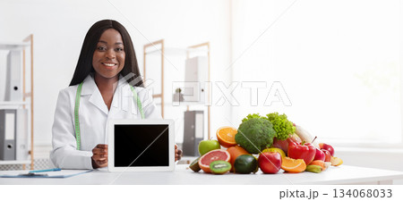Smiling afro woman nutritionist recommending application, showing empty digital tablet screen, clinic interior Smiling afro woman nutritionist recommending application, showing empty digital tablet screen, clinic interior 134068033