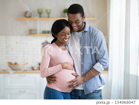 Black pregnant woman and her handsome husband embracing and smiling while spending time together near window at home. Romantic African American couple expecting new baby indoors. Parenthood concept 134068047