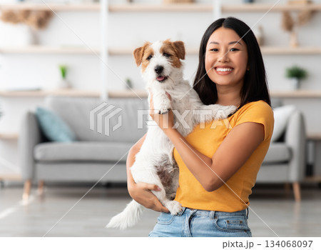 Portrait of cheerful young asian woman in casual outfit carrying her sweet fluffy dog jack russel terrier breed, pet and owner having good time together at home, living room interior, copy space 134068097