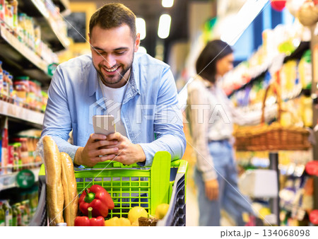 Smiling Male Customer Doing Grocery Shopping Using Smartphone Walking With Cart In Supermarket. Selective Focus. Man Using Groceries Shopping Application On Phone Bying Food In Super Market Smiling Male Customer Doing Grocery Shopping Using Smartphone Walking With Cart In Supermarket. Selective Focus. Man Using Groceries Shopping Application On Phone Bying Food In Super Market 134068098
