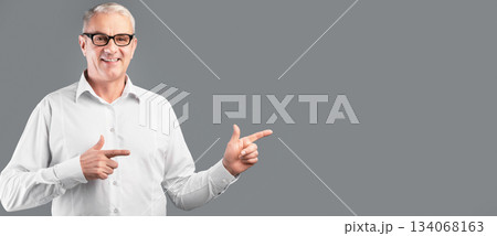Cool Offer. Smiling mature man in glasses pointing two index fingers aside at copyspace, gray studio wall, panorama 134068163