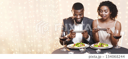 Young black couple sitting in restaurant and taking pictures of food with mobile phone, food bloggers celebrating Valentine's Day 134068244