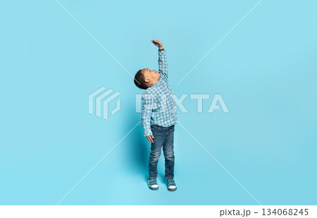 Little african american boy wishes he were taller, raising his hand to show how tall he wants to grow, child measuring his height, posing isolated on blue background, panorama, full length Little african american boy wishes he were taller, raising his hand to show how tall he wants to grow, child measuring his height, posing isolated on blue background, panorama, full length 134068245
