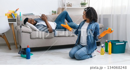 Domestic Duties Problem. Lazy black man relaxing on sofa with smartphone while his wife cleaning flat, displeased woman blaming husband, free space 134068248