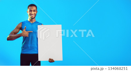 Cheerful Fitness Guy Holding Blank White Board For Text Pointing Finger At It Standing On Blue Background. Mockup, Studio Shot 134068251