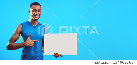 Workout Concept. Fit Man Holding Empty White Poster Pointing Finger At It Advertising Gym Over Blue Background. Studio Shot, Mockup 134068263
