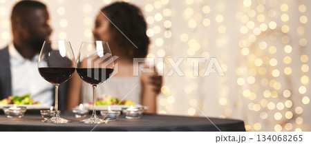 Selective focus on two glasses of wine over black couple in love, having festive dinner at restaurant Selective focus on two glasses of wine over black couple in love, having festive dinner at restaurant 134068265
