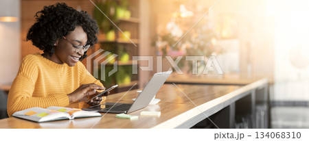 Happy black girl using mobile phone, working with laptop in cafe, side view, copy space 134068310