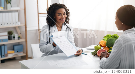Cheerful african woman dietician showing female patient medical chart, clinic interior, empty space 134068311