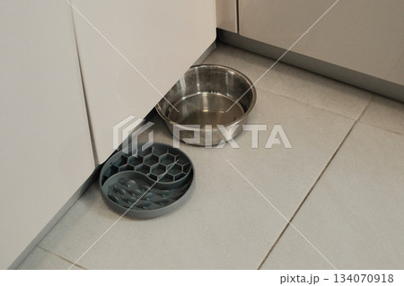A metal dog water bowl and a grey feeding pad sit on a tiled kitchen floor. The simple setup shows everyday pet care and home routine A metal dog water bowl and a grey feeding pad sit on a tiled kitchen floor. The simple setup shows everyday pet care and home routine 134070918