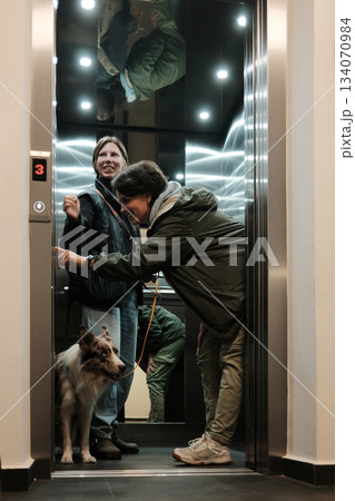 Two women stand in an elevator with a border collie on a leash. The scene reflects daily routine and the concept of urban living 134070984