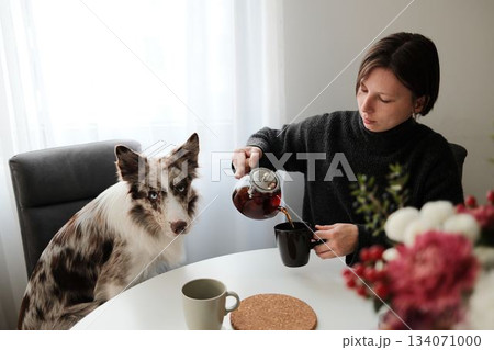 A woman pours tea at a breakfast table while her dog watches attentively, creating a warm domestic moment. The scene reflects companionship, calm routine, and quiet morning atmosphere A woman pours tea at a breakfast table while her dog watches attentively, creating a warm domestic moment. The scene reflects companionship, calm routine, and quiet morning atmosphere 134071000