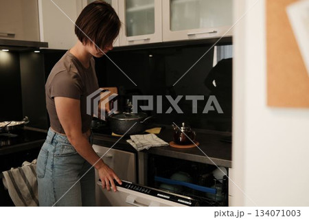 A woman loads a dishwasher while holding a cooking pot in a modern kitchen. The image conveys a simple household task within an everyday domestic routine A woman loads a dishwasher while holding a cooking pot in a modern kitchen. The image conveys a simple household task within an everyday domestic routine 134071003