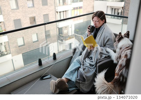 A woman wrapped in a blanket reads a notebook on a balcony while sipping a warm drink, as her dog stands nearby. The moment reflects calmness, comfort, and urban relaxation A woman wrapped in a blanket reads a notebook on a balcony while sipping a warm drink, as her dog stands nearby. The moment reflects calmness, comfort, and urban relaxation 134071020