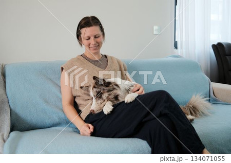A woman relaxes on a sofa holding her border collie on her lap. The scene shows calm bonding, trust, and a peaceful indoor moment with a pet 134071055