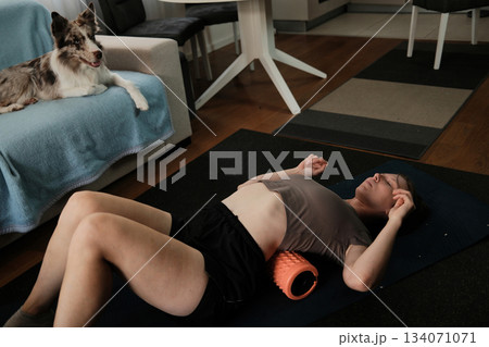 A woman lies on a foam roller while the border collie watches her from the sofa. The atmosphere shows home wellness with a peaceful pet presence A woman lies on a foam roller while the border collie watches her from the sofa. The atmosphere shows home wellness with a peaceful pet presence 134071071