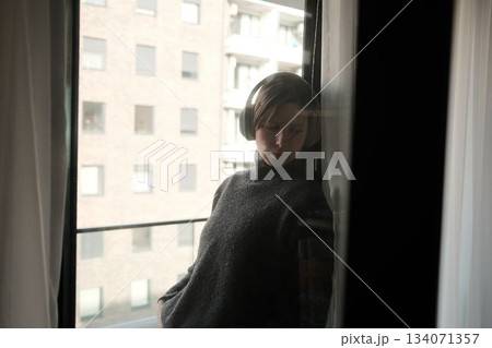 A woman in headphones leans near a window with a gentle reflection, creating a pensive and introspective scene 134071357